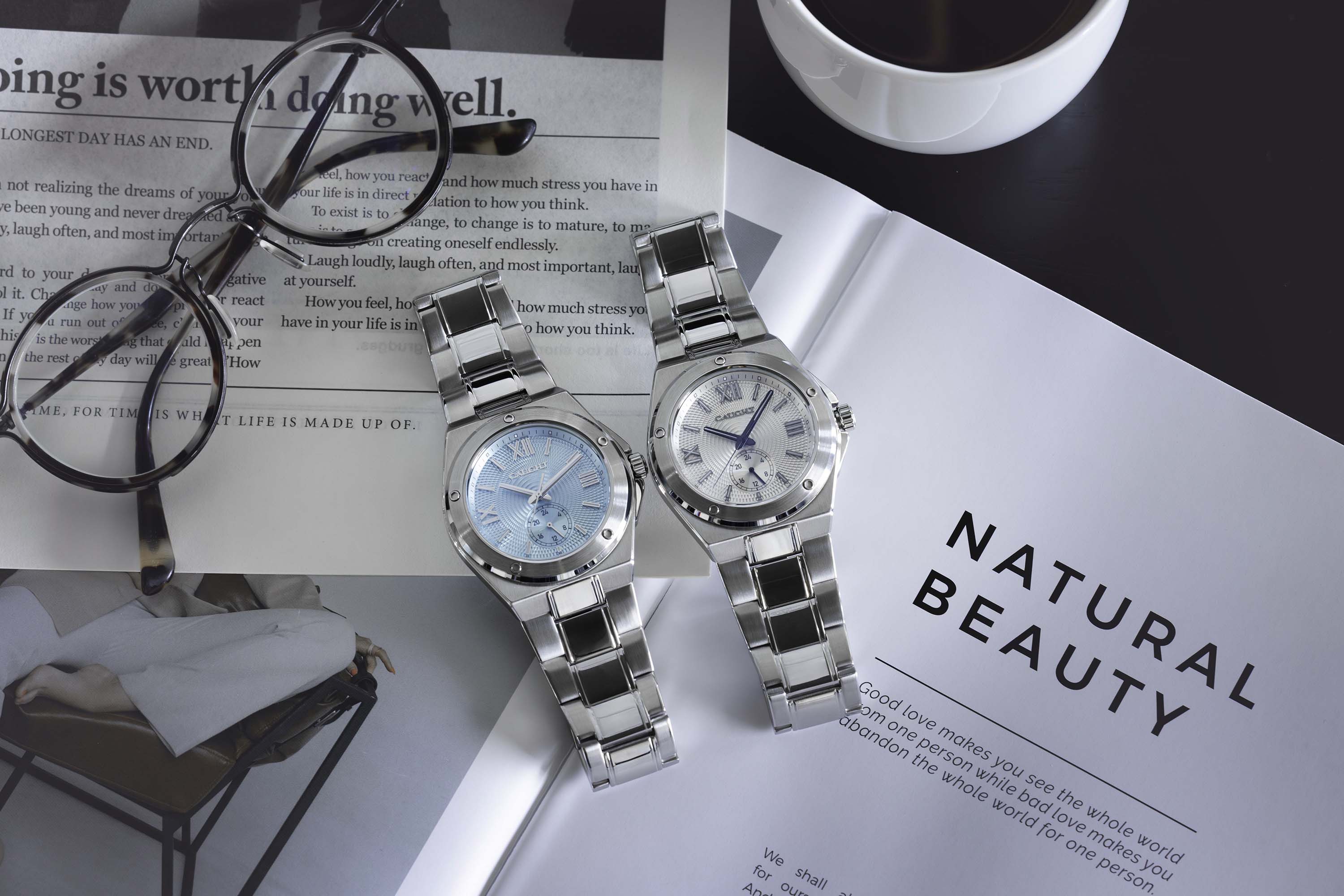 Two stainless steel watches on a magazine with a cup of coffee in the background